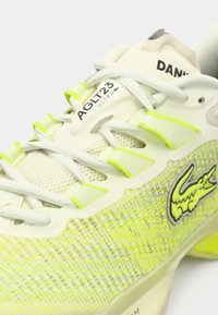 Lacoste AGLT23 Ultra sporta apaviem ir acs audums dzeltenzaļos toņos, pelēkas lentes, gumijas akcenti un izteiksmīgs logo.