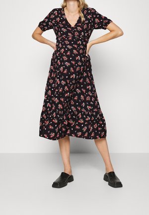 Frau trägt ein schwarzes Midikleid mit rot-weißem Blumenmuster, steht mit den Händen in den Hüften und schwarzen Slip-On-Schuhen.