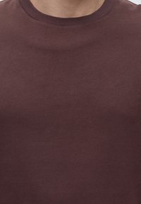 Gros plan d'une personne portant un t-shirt uni marron foncé à col rond, montrant le cou et le haut de la poitrine.