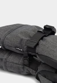 Zaino in tessuto grigio con spallacci neri regolabili e fibbie in plastica, che mostra da vicino i dettagli della chiusura e del materiale testurizzato.