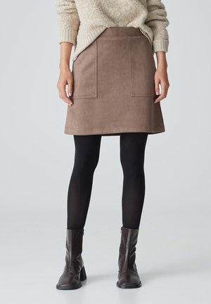 Brauer Wildlederrock mit zwei aufgesetzten Taschen, besäumtem Saum und einer leicht A-Linien-Form. Kombiniert mit schwarzen Leggings und braunen Stiefeletten.