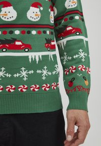 Grüner Strickpullover mit Schneemännern, roten Lastwagen mit Weihnachtsbäumen, Schneeflocken und Pfefferminzmustern in Rot, Weiß und Schwarz.