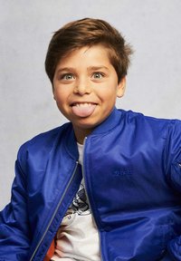 Giacca bomber blu con zip frontale, indossata sopra una t-shirt bianca con grafiche. Capelli corti castani, occhi verdi e espressione giocosa con la lingua di fuori.