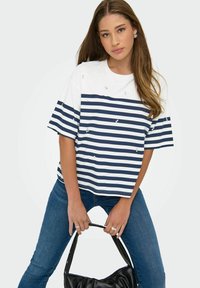 Jeune femme aux longs cheveux bruns portant une chemise striped blanche et marine avec de petites embellissements, un jean bleu, tenant un sac à main noir.