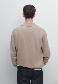 Man som står vänd mot vit vägg, klädd i beige tröja och mörka jeans med händerna avslappnat i fickorna.