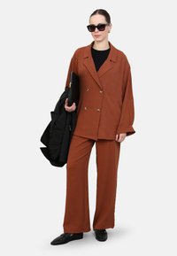 Blazer marron à double boutonnage et pantalon large en tissu texturé, associé à un haut noir, tenant une pochette noire et portant des lunettes de soleil.
