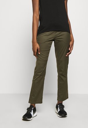 Osoba nosící olivově zelené kalhoty s rovnými nohavicemi, černé atletické boty s bílými podrážkami a černé tričko s krátkým rukávem na jednobarevném pozadí.