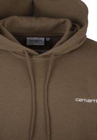 Bruine fleece hoodie met een trekkoordkap, met een witte "carhartt" logo op de borst en een zachte, gestructureerde stof. Maatlabel zichtbaar.