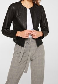 Chaqueta de cuero negra corta con escote redondo, diseño ajustado y dobladillo con volantes. Combinada con pantalones de cuadros gris claro de talle alto.