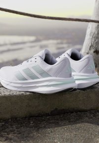 Paire de chaussures de course blanches Adidas avec des bandes gris clair reposant sur un rebord en pierre à l'extérieur, près d'un arbre et d'une barrière en corde.