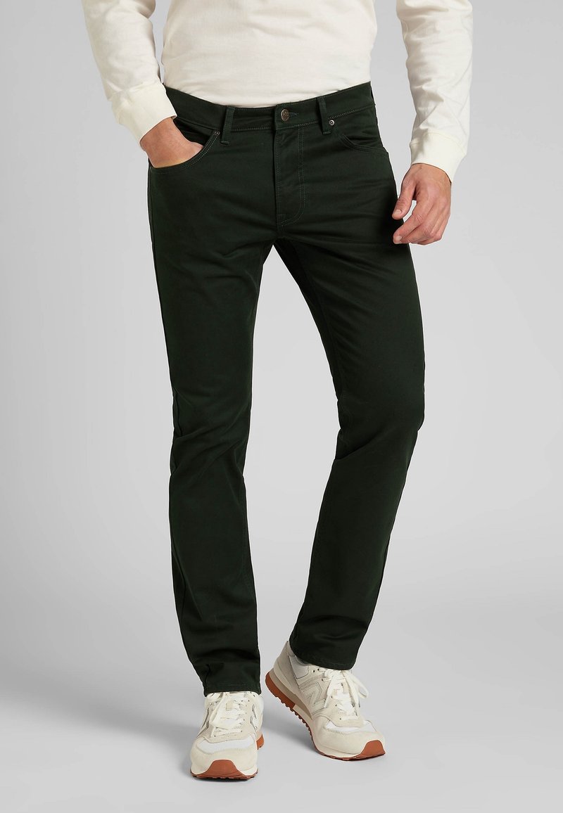 Homme portant un pantalon slim noir, des baskets blanches et une chemise crème à manches longues, debout avec une main dans la poche.