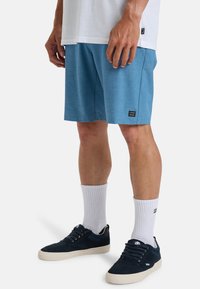 Blauwe sportshorts van gestructureerde stof, met een zwart logo op de linker bovenbeen, gecombineerd met witte crew-sokken en donkerblauwe sneakers.