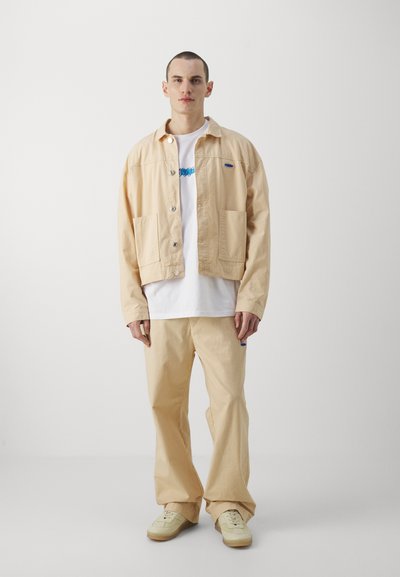 Veste en coton beige avec deux poches avant, fermeture à boutons, associée à un t-shirt blanc et un pantalon beige assorti, ainsi qu'à des baskets de couleur claire.