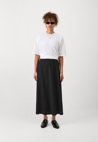 Črna maksi krila z elastičnim pasom, v kombinaciji s sproščenim belim t-shirtom. Dodatki vključujejo plastične ogrlice in temne sončne očala.