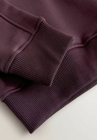 Gros plan sur une manche de sweat-shirt violet foncé avec poignets et ourlet côtelés sur une surface claire.