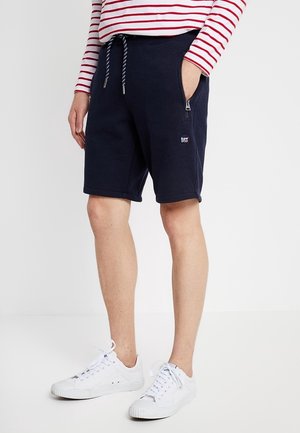 Homme portant un short bleu marine à cordon avec poche zippée, des baskets blanches, et un t-shirt à manches longues rayé rouge et blanc, debout sur un sol blanc.