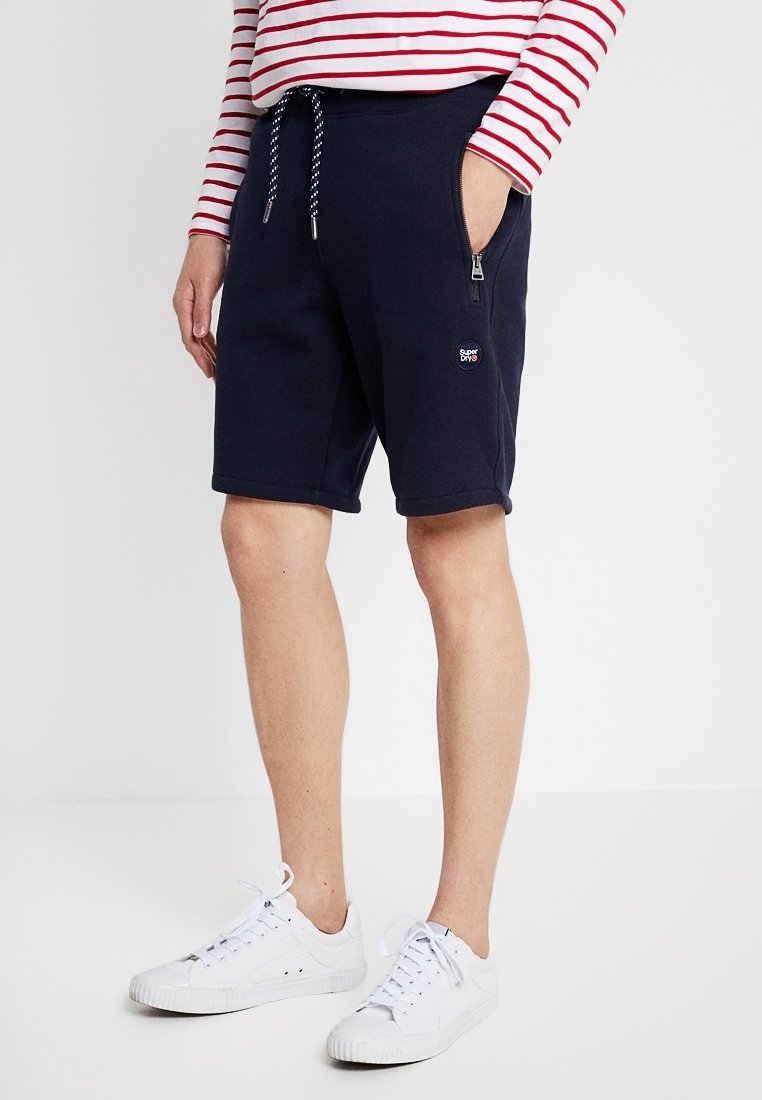 Homme portant un short bleu marine à cordon avec poche zippée, des baskets blanches, et un t-shirt à manches longues rayé rouge et blanc, debout sur un sol blanc.