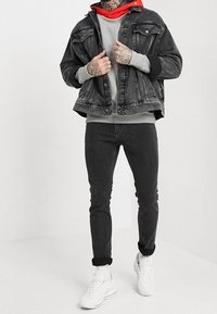 Svart denimjacka, grå sweatshirt med röd huva, svarta slim-fit jeans och vita sneakers. Anmärkningsvärda tatueringar på händerna.