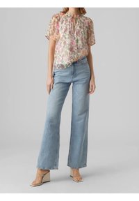 Blouse fleurie transparente à col noué, avec des motifs roses, verts et orange, associée à un jean large bleu clair et des sandales transparentes.