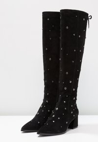 Mustat mokkanahkaiset polvipituiset saappaat, joissa on paksut korot, kärjet ovat terävät ja joissa on hajallaan hopeanvärisiä niittejä ja koristeellisia yksityiskohtia.