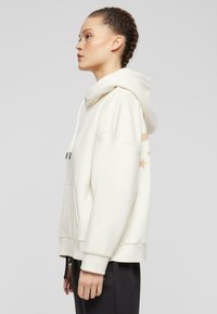 Cream hoodie with a kangaroo pocket, drawstring hood, and decorative stars pattern on the sleeve. Material appears soft and textured.