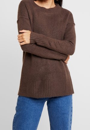 Person wearing a brown knit sweater with ribbed cuffs and hem, paired with blue denim jeans, arms crossed at the waist.