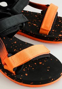 Čierne a oranžové sandále majú nastaviteľné popruhy, textúrovanú podrážku a škvrnitý vzor na čiernom základe s oranžovým vonkajším okrajom.
