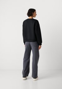 Donna con capelli corti e ricci sta in piedi di spalle indossando una felpa nera, pantaloni larghi grigi, orecchini a cerchio e sneakers bianche su uno sfondo neutro.