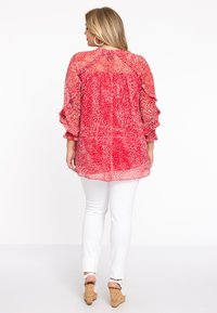 Langärmlige rote Bluse mit weißem abstraktem Muster, Rüschensäumen, leichtem Stoff, kombiniert mit weißen Hosen und Wedgesandalen.