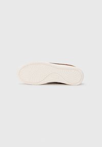 Braune Sneaker mit einer Gummisohle, die ein Wellenmuster aufweist, einem strukturierten Obermaterial und einer kontrastierenden weißen Zwischensohle.