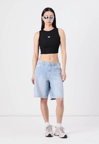 Svart cropped tank topp med et lite hjerteemblem, kombinert med lyseblå denimshorts. Modellen har på seg sølvfargede solbriller og lyse sneakers.