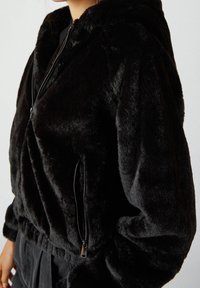 Veste bomber en fausse fourrure noire avec capuche, dotée d'une fermeture éclair à l'avant et d'un cordon réglable à l'ourlet. Texture douce et pelucheuse.