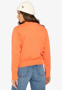 Oranje sweatshirt met een ronde halslijn, geribbelde manchetten en zoom. De stof is zacht met een gladde textuur, gecombineerd met blauwe spijkerbroek.
