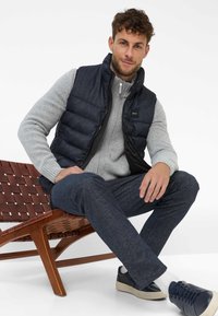 Marine puffer gilet over een grijze gebreide trui, gecombineerd met gestructureerde donkere broek en marineblauwe sneakers. De setting bevat een houten stoel.