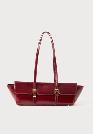 Burgundy nahkakäsilaukku, jossa on kaksi hihnaa, jämäkkä muoto ja kaksi etutaskua, jotka on kiinnitetty kultaisilla soljilla. Sujuva pinta ja viisto sivu.