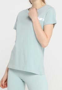 Žena nosící světle modrý top s krátkým rukávem a odpovídající legíny, se reflektivním pruhem na hrudi, pózující na bílém pozadí.