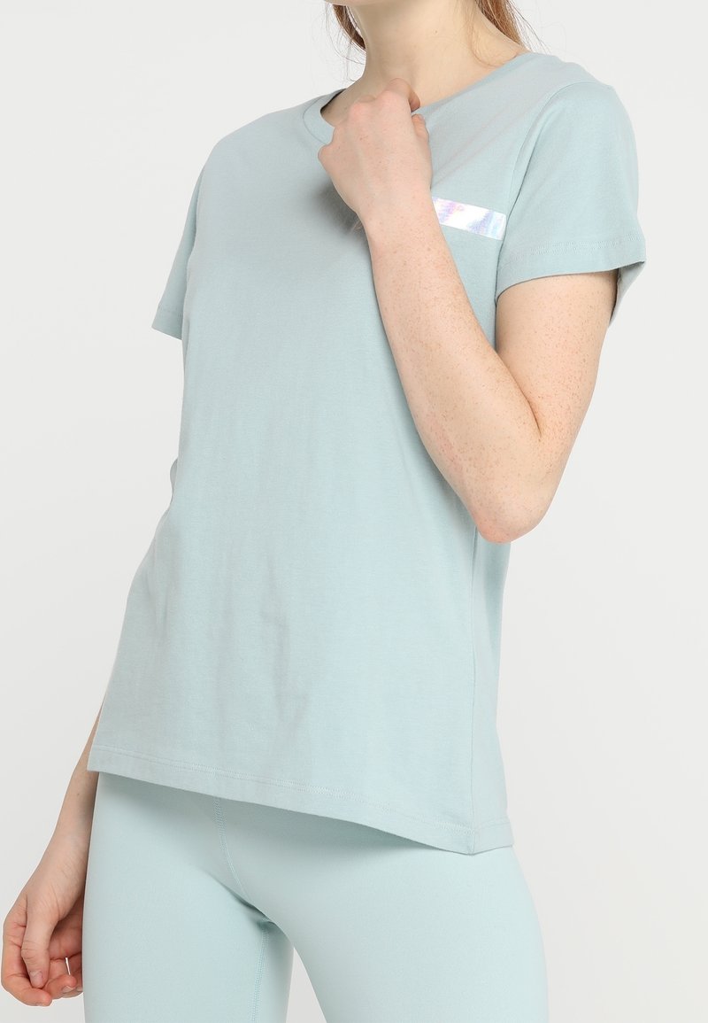 Žena nosící světle modrý top s krátkým rukávem a odpovídající legíny, se reflektivním pruhem na hrudi, pózující na bílém pozadí.