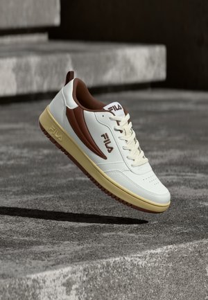 Valge ja pruun FILA toss kreemja tallaga, õhus hallide betoonastmete kohal, all varju heites.