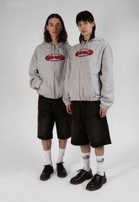 Des sweats à capuche gris zippés avec un logo rouge, associés à des shorts en denim noir. Tous deux portent des chaussures noires et des chaussettes blanches avec des accents imprimés.