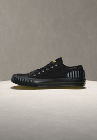Baskets basses noires avec semelle en caoutchouc texturée et bout renforcé, lacets noirs et détail subtil du logo jaune sur la languette et le talon.