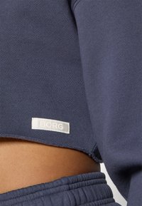 Marineblå cropped sweatshirt med teksturert stoff, rå kant og en hvit "BORG"-lapp sydd på bunnen.