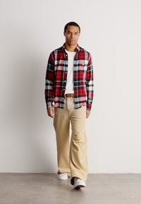 Hombre caminando en interiores con una camisa de cuadros roja y negra, camiseta blanca, pantalones beige, cinturón marrón y zapatillas blancas sobre un fondo liso.