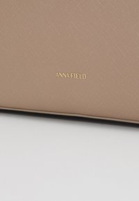 Gros plan d'une surface texturée beige avec le nom de marque ANNA FIELD embossé en or, centré près du bas.
