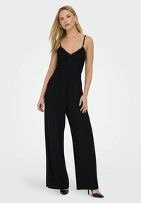 Combinaison noire avec un corsage en dentelle, des bretelles spaghetti, un design à jambes larges et des accents texturés, associée à des talons hauts noirs.