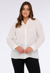 Weiße Bluse mit langen Ärmeln und Knopfleiste, die mit kleinen Punkten und kontrastierenden Bündchen versehen ist. Der Stoff wirkt leicht und glatt.