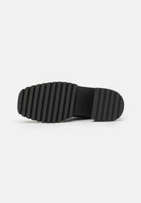Semelle de chaussure noire avec de larges rainures horizontales pour l'adhérence, présentée à plat sur fond blanc.