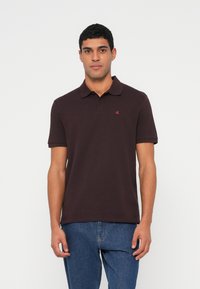 Tmavě hněré polo tričko s krátkými rukávy a límečkem. Na levé straně hrudi je malé červené logo. Kombinováno s modrými džínami z denimu.