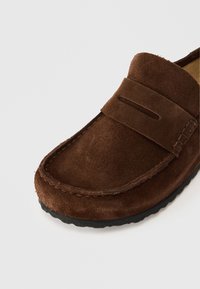 Καφέ γυναικείο μοκασίνι από καστόρι με στρογγυλερή μύτη, διακοσμημένο με ραφτή λεπτομέρεια και επίπεδη λαστιχένια σόλα για καλύτερη πρόσφυση. Το επάνω μέρος έχει λεία υφή.