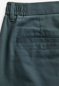 Pantaloni di colore verde scuro, con una texture liscia, cintura elastica e una tasca posteriore singola con apertura orizzontale.