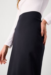 Femme portant une jupe noire ajustée et un haut blanc à manches longues, debout avec une main touchant le côté de la jupe.